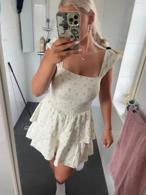 Vit blommig kortklänning med volang - Supersöt vit kortklänning med små blommor och volangdetaljer. Klänningen har smockad överdel, breda axelband och en luftig kjol med dubbla lager. Perfekt för varma dagar och ger en romantisk vibe.
