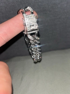 Iced ut silverfärgat kedjearmband - Säljer ett glittrande silverfärgat kedjearmband med massor av små, gnistrande stenar över hela länkarna och låset. Armbandet har en chunky design och ett fyrkantigt lås täckt av stenar. Perfekt för dig som gillar att sticka ut med accessoarer.