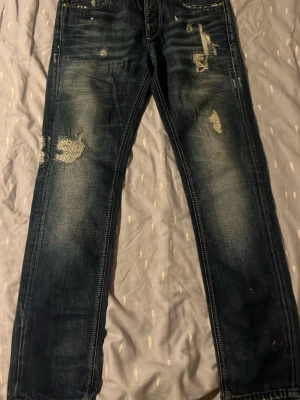 Mörkblå slitna jeans med patchar - Säljer ett par mörkblå jeans med slitningar och patchade detaljer på både fram- och baksida. Jeansen har raka ben och klassisk femficksmodell. Materialet är denim och de har en cool, använd look som passar streetwear-stilen.