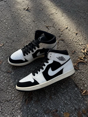 Svarta och vita Air Jordan 1 Mid - Snygga Air Jordan 1 Mid sneakers i svart och vitt läder med klassisk Nike Swoosh på sidorna och Jordan Wings-logga vid ankeln. Skorna har svart snörning och perforerad tå för extra ventilation. Perfekta för dig som gillar streetwear och ikonisk design.