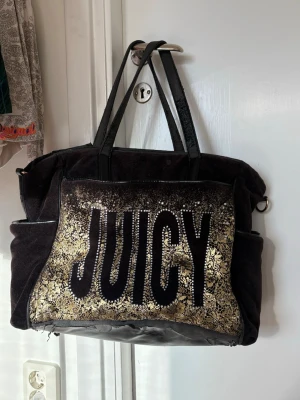Juicy Couture svart handväska med paljetter - Stor svart handväska från Juicy Couture med glittrande guldpaljetter och spetsdetaljer på framsidan. Tydlig JUICY-logga i svart sammet. Väskan har dubbla handtag och sidofickor, tillverkad i mjukt tyg och konstläder. Den är sliten så därav priset!🌼