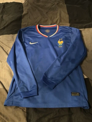Blå långärmad Frankrike Nike tröja - Snygg blå långärmad fotbollströja från Nike med Frankrikes landslagsmärke och två stjärnor på bröstet. V-ringad krage med röd, vit och blå kant. Tillverkad i ett lätt och ventilerande material, perfekt för träning eller match.