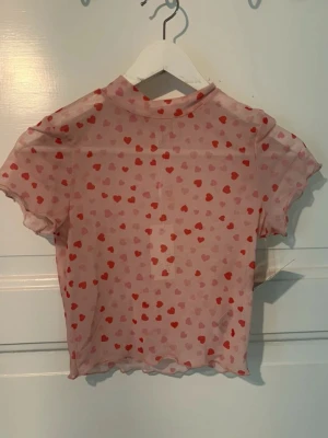 Rosa mesh topp med hjärtan - Söt rosa mesh topp med röda och ljusrosa hjärtan över hela tyget. Toppen har korta ärmar, hög hals och vågiga kanter. Perfekt för dig som vill ha en lekfull och trendig look.