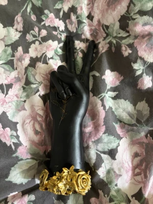 Svart handformad smyckeshållare - Unik smyckeshållare i form av en svart hand som gör peace-tecknet. Basen är dekorerad med detaljerade guldfärgade blommor. Perfekt för att förvara ringar, armband eller andra små accessoarer. Nypris ca 299kr