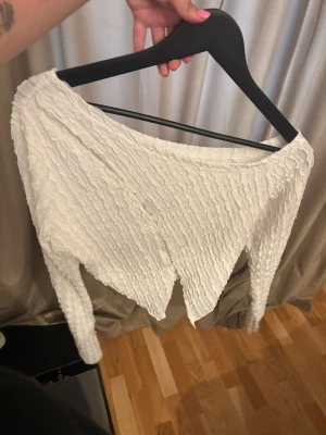 Vit långärmad topp med struktur - Säljer en vit långärmad topp med unik strukturerad yta och asymmetrisk nederkant. Toppen har en offshoulder-design och är tillverkad i ett stretchigt syntetmaterial. Perfekt för dig som vill sticka ut med en cool och modern look.