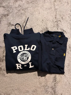 Mörkblå Ralph Lauren hoodie och Ralph Lauren piké - Mörkblå hoodie från Polo Ralph Lauren med vit tryckt logga och tigrar på bröstet. Klassisk huva med dragsko och ribbade muddar. Perfekt för en avslappnad och sportig look. Begagnad så saknar därför kvitto men tros vara äkta. Ingen av plaggen har inga slitningar. Hoodien är XXL och piken XL.