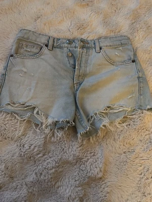Ljusblå jeansshorts med fransig kant - Säljer ett par ljusblå jeansshorts med fransig, rå kant och slitna detaljer. Shortsen har klassisk femficksdesign, knappgylf och en loose fit som ger en avslappnad vibe. Perfekta för sommardagar och festivaler.
