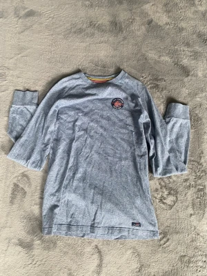 Grå/blå långärmad t-shirt med patch - Säljer en grå/blå långärmad t-shirt med rund hals och en liten patch med tryck på bröstet. T-shirten har breda muddar vid ärmsluten och är tillverkad i mjuk bomull. Perfekt för en avslappnad stil.