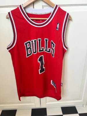 Chicago Bulls basketlinne röd - Rött basketlinne med Chicago Bulls-tryck och nummer 1 på framsidan. Klassisk design med svarta och vita detaljer runt hals och ärmhål. Tillverkat i ventilerande meshmaterial, perfekt för basket eller träning.