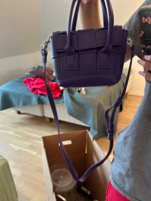Mini lila väska  - Söt väska från nakd.Inga defekter då den knappt är använd,säljer för att ingen använder den.Den har ett lika räfflande mönster och två små handtag och ett stort så du kan hänga väskan runt dig🥰ställ frågor 