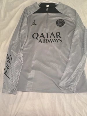 Grå PSG träningströja från Jordan - Snygg grå långärmad träningströja från PSG och Jordan med halv dragkedja, svart logga på bröstet och 'Qatar Airways' tryck framtill. Ärmarna har diskret mönster och texten 'Paris' tryckt på sidan. Tillverkad i lätt och ventilerande funktionsmaterial.