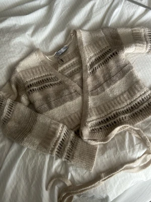 Beige stickad omlottkofta från Zara - Säljer en beige stickad omlottkofta från Zara med breda ränder i olika nyanser av beige och brunt. Koftan har långa ärmar, ribbade muddar och knytband i midjan. Materialet är mjukt och fluffigt, perfekt för lager på lager och en chill look.