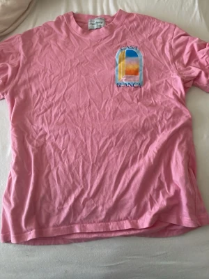 Rosa Casa Blanca t-shirt med tryck - Säljer en rosa t-shirt från Casa Blanca med färgglatt grafiskt tryck fram och bak. T-shirten har rund hals och korta ärmar. Materialet är mjuk bomull och trycket har inslag av blått, gult och orange. Perfekt för dig som gillar statement-plagg.