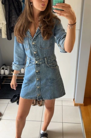 Kort jeansklänning med knappar - Säljer en snygg kort jeansklänning i blå denim från Zara med långa ärmar och knappar hela vägen framtill. ANVÄND EN GÅNG! Klänningen har fickor på bröstet och vid höfterna samt bältdetaljer i midjan. Perfekt för dig som gillar en avslappnad men trendig stil. 