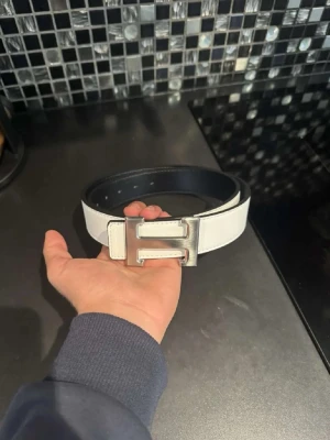 Vitt skärp med silverfärgat H-spänne - Snyggt vitt bälte med ett stort, silverfärgat H-format spänne. Bältet har en stilren design och passar perfekt till jeans eller byxor för en clean look. Insidan är svart vilket ger en cool kontrast. Perfekt accessoar för att lyfta din outfit.