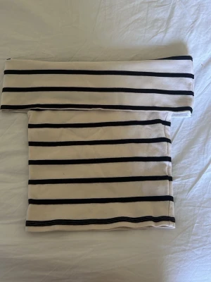 Randig offshoulder topp Gina Tricot - Säljer en vit offshoulder topp från Gina Tricot med svarta horisontella ränder. Toppen är axelbandslös och har en tight passform, perfekt för en trendig och stilren look. Materialet känns mjukt och stretchigt.