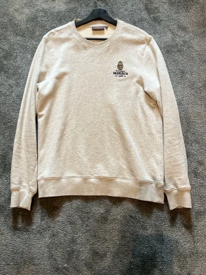 Ljusgrå sweatshirt från Morris - Ljusgrå sweatshirt från Morris med broderad logga och emblem på bröstet. Tröjan har rund halsringning, ribbade muddar vid ärmslut och nederkant samt en klassisk, avslappnad passform. Perfekt för en stilren och enkel look.