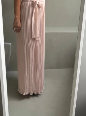 Ljusrosa plisserad byxdress med knyt - Säljer en ljusrosa byxdress med plisserad design och ett brett knytband i midjan. Den har en mjuk och flowy känsla, perfekt för dig som gillar en feminin och elegant stil. Passar till både sneaker/högklackat el sandaler.