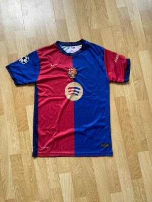 FC Barcelona matchtröja Nike - Snygg FC Barcelona matchtröja från Nike i röd och blå med klubbmärke, Spotify-logga och Champions League-märke på ärmen. Tröjan har Lamine Yamal och nummer 19 på ryggen samt UNHCR-logga. Tillverkad i lätt och ventilerande polyester, perfekt för fotboll.