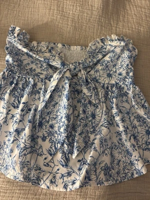 Blommig axelbandslös topp med rosett - Söt axelbandslös topp med blått blommönster på vit botten och en stor rosett framtill. Toppen har volangdetaljer upptill och är perfekt för varma dagar. Materialet känns lätt och luftigt.