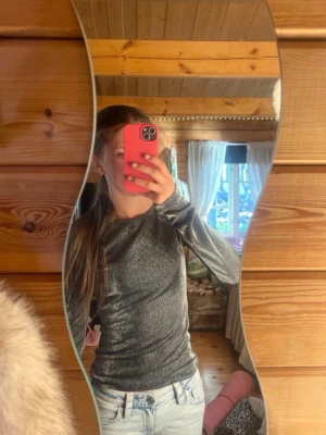 Glittrig långärmad topp i silver - Supersnygg långärmad topp i glittrigt silvermaterial. Figurnära passform och rund halsringning, perfekt för dig som vill sticka ut lite extra. Passar till både jeans och kjol för en cool look.