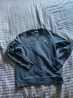 Mörkblå sweatshirt från Morris - Mörkblå sweatshirt från Morris med rund halsringning och ribbade muddar. Tröjan har en liten vit broderad logga på bröstet och klassisk college-look. Perfekt till jeans eller chinos för en avslappnad stil.