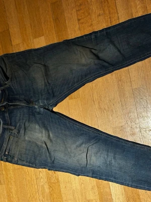 Mörkblå raka jeans med slitningar - Säljer ett par mörkblå jeans med raka ben och snygga slitningar på framsidan. Klassisk femficksmodell med knappgylf och kontrastsömmar. Jeansen har en avslappnad passform och är tillverkade i bomull. Perfekta för en cool och avslappnad stil.