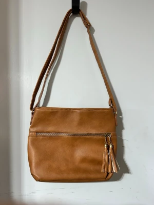 Kamelbrun axelväska med tofsdetalj - Snygg kamelbrun axelväska i slätt konstläder med justerbar axelrem. Väskan har en dragkedjeficka framtill och dekorativa tofsar i matchande färg. Enkel, stilren design som passar till många outfits.