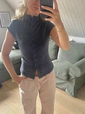 Mörkblå figurnära blus med knappar - Snygg mörkblå blus med hög krage och knappar hela vägen framtill. Modellen är figurnära och har korta ärmar. Perfekt till jeans eller kjol för en trendig look. Materialet ser ut att vara mjukt och stretchigt.