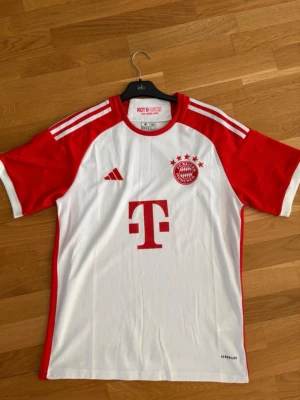 Bayern München matchtröjor Adidas - Två snygga Bayern München matchtröjor från Adidas. Den ena är vit med röda ärmar och detaljer, klassisk klubbmärke och T-Mobile-logga. Den andra är helvit med röda ränder på axlarna, krage och klubbmärke. Båda i funktionsmaterial, perfekta för fotboll eller träning.