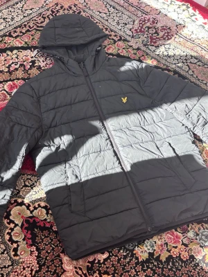 Svart pufferjacka från Lyle & Scott - Säljer en svart pufferjacka från Lyle & Scott med gul broderad logga på bröstet. Jackan har huva, dragkedja framtill och två sidofickor. Perfekt för kalla dagar och riktigt skön att bära. Materialet är syntet och jackan är quiltad för extra värme. Dragkedjan är lite trasig där nere men går absolut att fixa! Det finns en vit fläck på luvan också, och det är dom två är jag skulle säga dom enda små skador på jackan! Möts gärna i Stockholm!