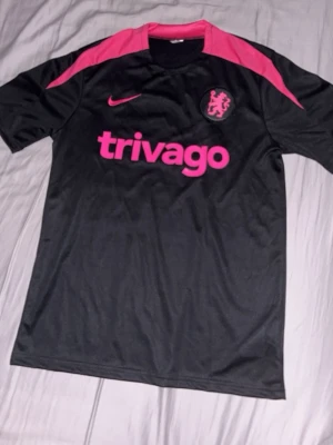 Chelsea Nike svart träningsset - Säljer ett svart Chelsea träningsset från Nike med rosa detaljer. Tröjan har rosa logga, klubbmärke och 'trivago' tryck på bröstet. Shortsen har matchande rosa Nike-logga och Chelsea-märke. Tillverkad i lätt och ventilerande material, perfekt för träning.
