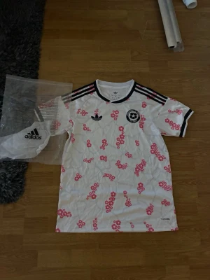 Chile Bortaställ - Vit fotbollströja från Adidas med svarta detaljer och rosa blommönster. Tröjan har svarta ränder på axlarna, svart krage och tryckt märke för Federación de Fútbol de Chile på bröstet. Perfekt för dig som gillar unika sportkläder.