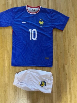Frankrike fotbollströja Mbappé #10 Nike - Säljer en blå Frankrike fotbollströja från Nike med Mbappé och nummer 10 på ryggen. Tröjan har vita detaljer, röd krage och landslagets emblem på bröstet. Tillhörande vita shorts med emblem ingår. Perfekt för match eller träning.
