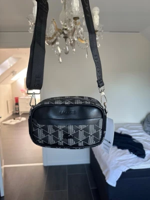 Svart axelväska från Lacoste - Snygg svart axelväska från Lacoste med vitt geometriskt mönster och justerbar axelrem med logga. Väskan har en dragkedja upptill, ett ytterfack och detaljer i svart konstläder. Perfekt för att bära det viktigaste med stil.