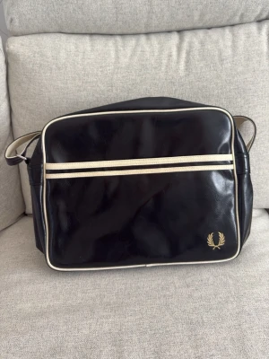 Svart axelväska från Fred Perry - Snygg svart axelväska från Fred Perry i konstläder med vita detaljer och logga i hörnet. Väskan har justerbar axelrem, dragkedja upptill och ett stort ytterfack med dragkedja. Perfekt för att bära allt du behöver till skolan eller stan.