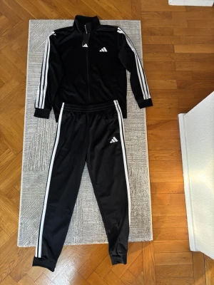 Svart Adidas träningsset  - Säljer ett svart träningsset från Adidas med klassiska vita ränder längs ärmar och ben. Setet består av en zip-jacka och byxor, båda med Adidas-logga på bröst och lår. Tillverkat i ett mjukt och stretchigt material, perfekt för träning eller chill. Helt nytt med prislapp kvar på. 