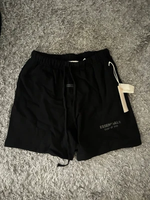 Svarta shorts Essentials Fear of God - Svarta shorts från Essentials Fear of God med elastisk midja och snörning. Klassisk loose fit och diskret logga på vänster ben. Perfekta för chill dagar eller streetwear-stil. Tillverkade i mjuk bomull för maximal komfort.