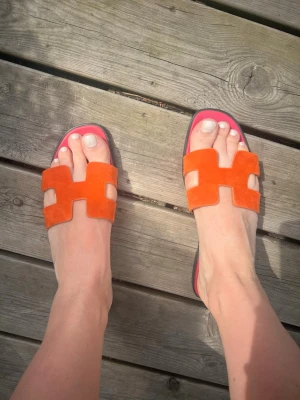 Orangeröda sandaler i mocka - Säljer ett par trendiga sandaler med orangeröda mockarem och rosa innersula. De har en öppen tå och breda remmar i ett H-format snitt. Perfekta för sommaren och riktigt snygga till jeans eller klänning.