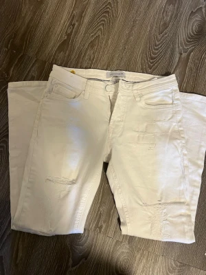 Vita distressed jeans från Revolution - Säljer ett par vita jeans från Revolution med snygga slitningar och femficksmodell. Jeansen har raka ben och klassisk knappgylf. Materialet är bomull med lite stretch för extra komfort. Perfekta för dig som gillar en clean men edgy stil. Skriv för fler frågor och bilder