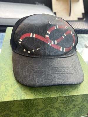 Svart keps med ormtryck från Gucci - (100% ÄKTA!) Säljer en svart keps från Gucci med ikoniskt ormtryck i rött, vitt och svart på framsidan. Kepsen har mesh på sidorna och justerbart band bak med röd och blå rand. Perfekt för dig som vill ha en statement-accessoar. Pris går att diskutera vid snabb affär!