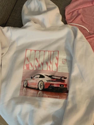 Vit hoodie med Porsche-tryck - Snygg vit hoodie med stor grafisk Porsche-bil på ryggen och rosa Porsche-logga på bröstet. Hoodien har huva med dragsko och en klassisk känguruficka framtill. Perfekt för dig som gillar bilar och streetwear. Materialet känns mjukt och bekvämt.