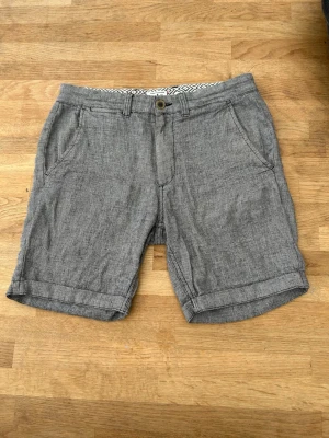 Grå linneshorts med fickor - Snygga grå shorts i linne med avslappnad passform och uppvikta benslut. Shortsen har framfickor, bakfickor med dragkedja och bälteshällor. Perfekta för varma dagar och enkel att matcha med olika stilar.