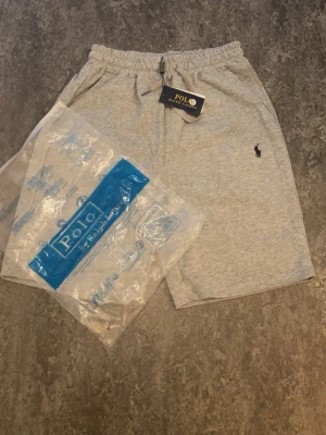 Grå shorts från Polo Ralph Lauren - Snygga ljusgrå shorts från Polo Ralph Lauren med elastisk midja och dragsko. Klassisk broderad logga på vänster ben. Perfekta för sommaren och tillverkade i mjuk bomull för maximal komfort.