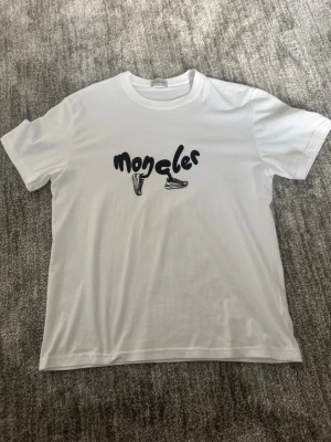 Vit t-shirt med tryck - Vit t-shirt med svart tryck på bröstet där det står 'moncler' och två sneakers illustrerade. Klassisk rund halsringning och korta ärmar. Tillverkad i mjuk bomull för en skön känsla och enkel stil.
