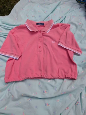 Rosa croppad pikétröja Fred Perry - Säljer en rosa croppad pikétröja från Fred Perry med vit logga på bröstet. Tröjan har klassisk krage, tre knappar framtill och vita ränder på krage och ärmslut. Perfekt för dig som gillar sportig och trendig stil. Tröjan är croppad av mig och har resårband ner till
