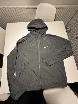 Nike Miler Grå Vindjacka - Snygg grå vindjacka från Nike med huva och dragkedja framtill. Jackan har två sidofickor med dragkedja och en vit Nike-logga på bröstet. Tillverkad i lätt och vindtätt material, perfekt för blåsiga dagar.
