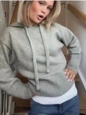 Ljusgrå stickad hoodie - Mysig ljusgrå stickad hoodie med huva och dragsko. Tröjan har bred ribbstickad mudd vid ärmslut och nederkant, vilket ger en schysst passform. Perfekt för dig som gillar en avslappnad och trendig stil. Materialet ser ut att vara mjukt och värmande ullblandning.
