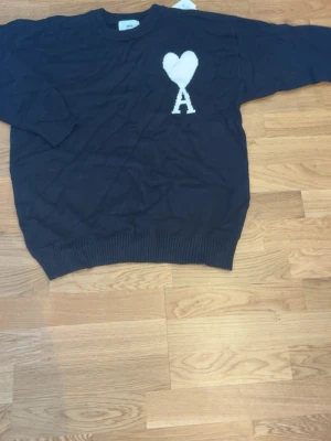 Svart stickad tröja från Ami Paris - Säljer en svart stickad tröja från Ami Paris med ett vitt hjärta och bokstaven A broderat på bröstet. Tröjan har rund hals, långa ärmar och ribbade muddar vid ärmslut och nederkant. Perfekt för dig som gillar stilrena detaljer och streetwear vibes.