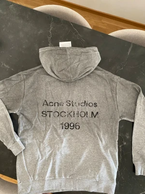 Grå hoodie från Acne Studios - Säljer en grå hoodie från Acne Studios med trycket 'Acne Studios STOCKHOLM 1996' på ryggen. Klassisk passform med huva och ribbade muddar vid ärmslut och nederkant. Perfekt för en chill och stilren look.